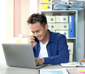 Man sitting at a desk working on a laptop while holding a phone to his ear