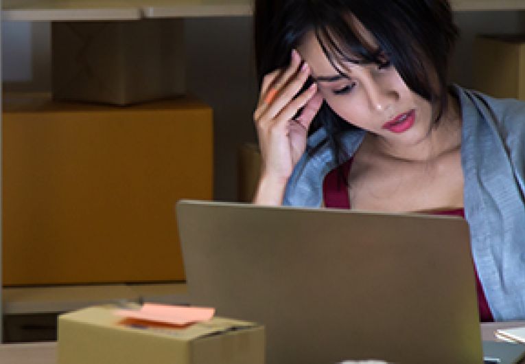 Woman holding her head while looking at a laptop with boxes on shelves behind her.